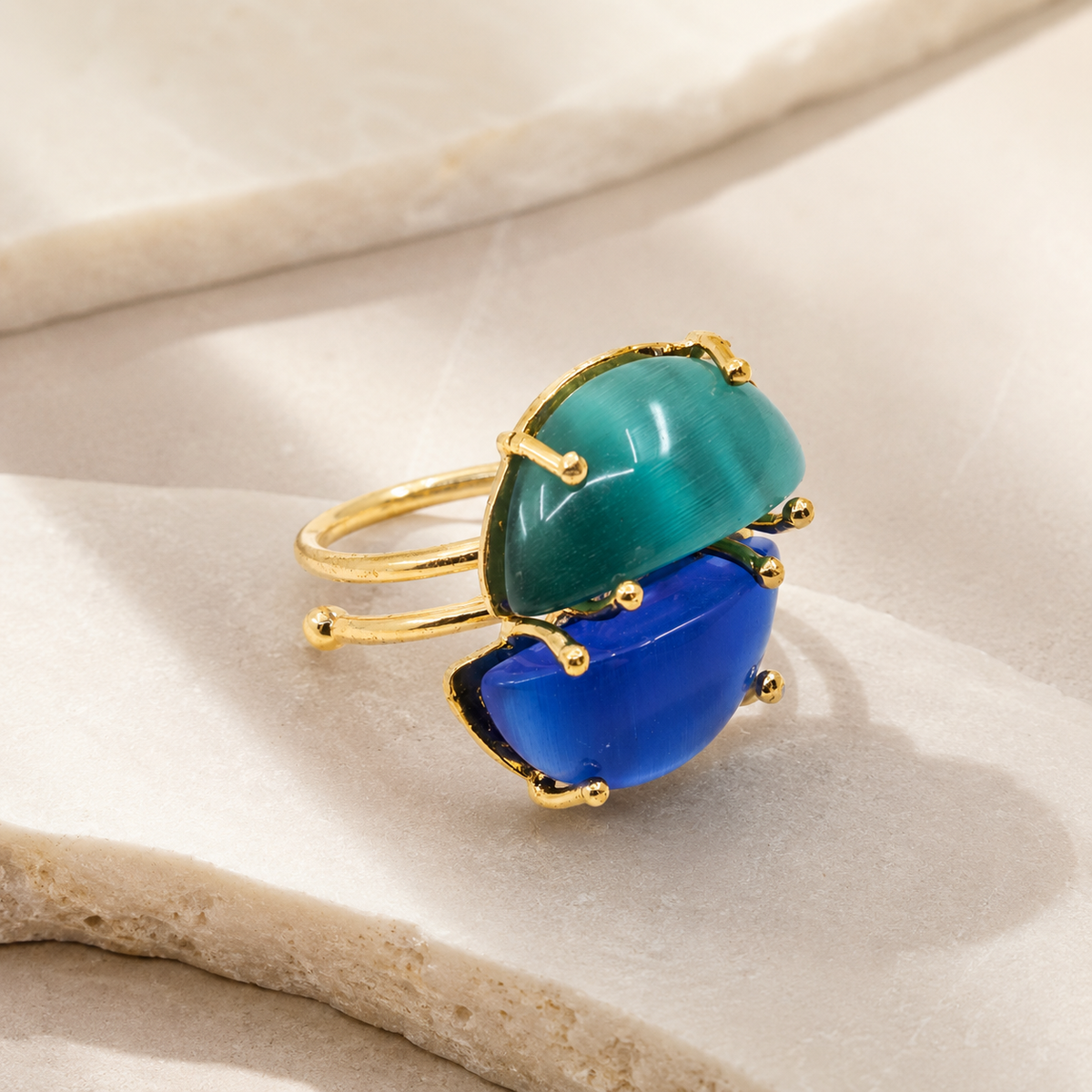Angled view of a gold-plated adjustable ring with two cat-eye style stones in blue and teal.