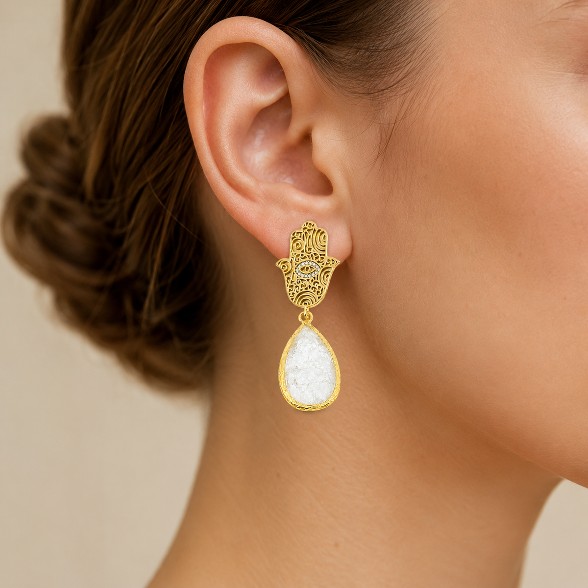 Gold hamsa hand drop earrings with white teardrop stones on a white background.