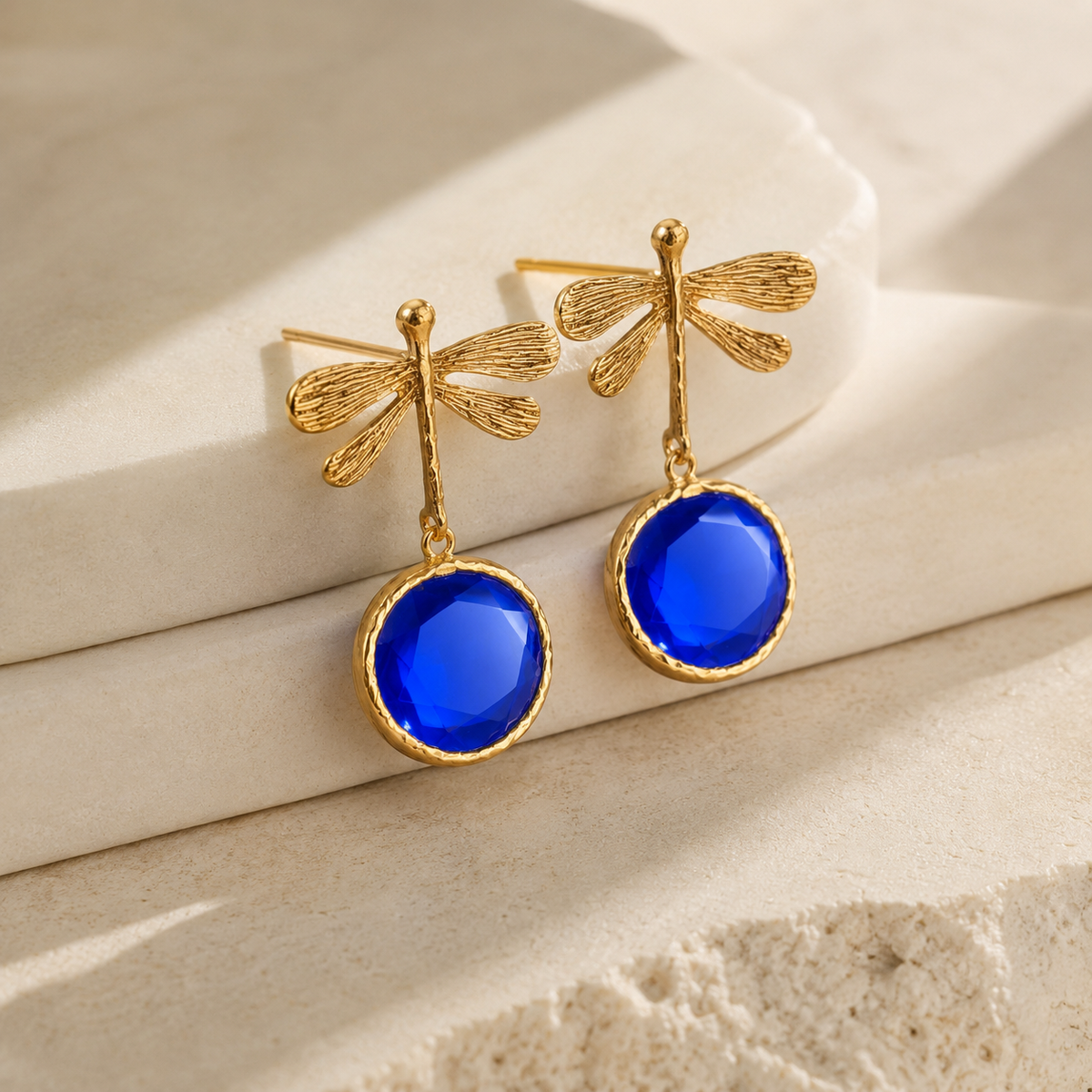 Angle view of dragonfly earrings with faceted blue drops and gold-plated details.