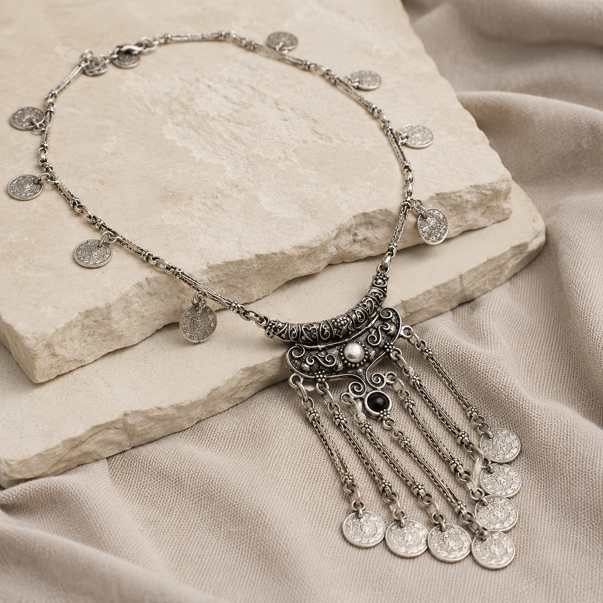 Angled view of a silver-tone coin pendant necklace with tassel drops and black stone detail.