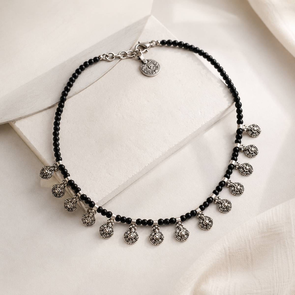Angled view of a black bead necklace with oval charm drops and silver-tone details.
