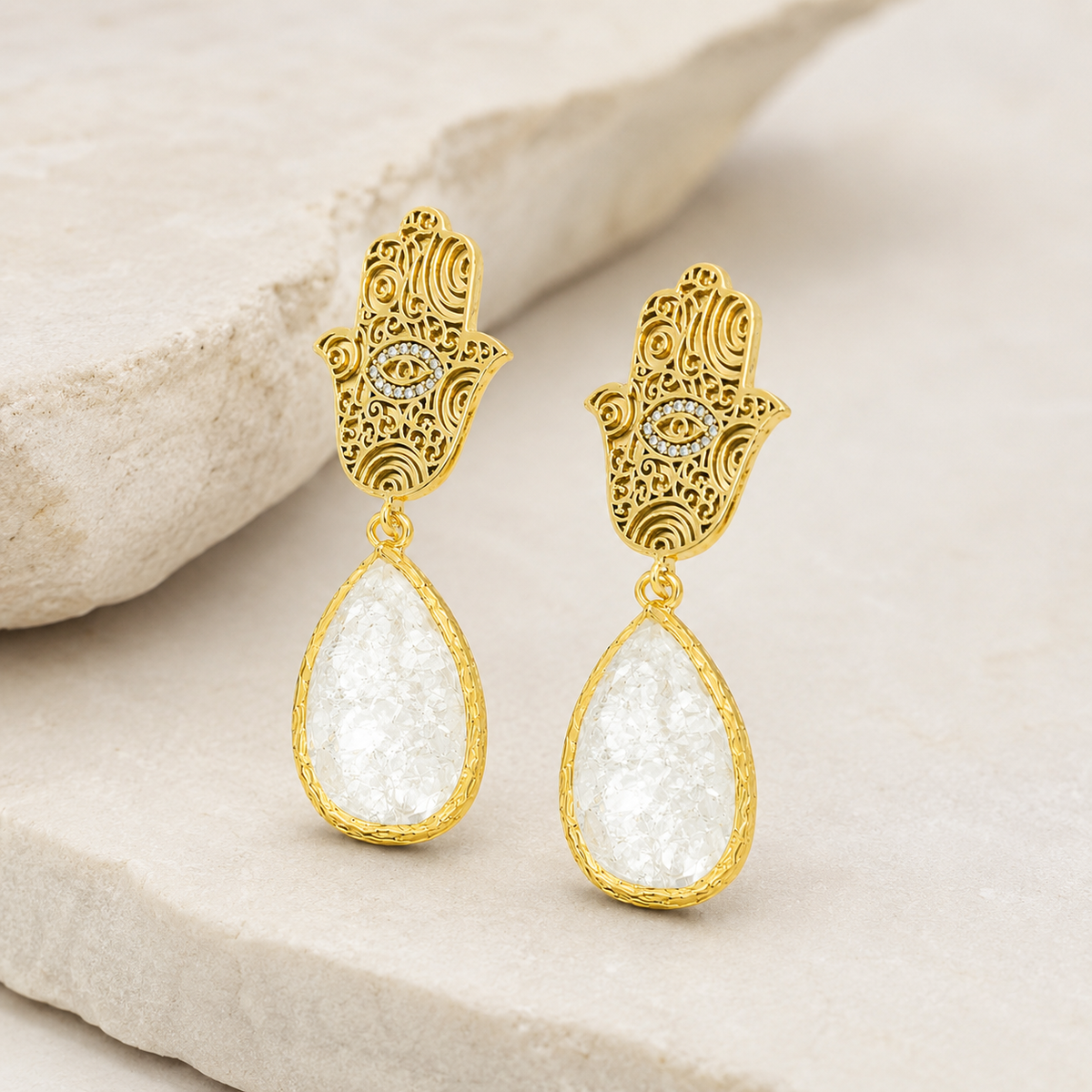 Angle view of gold hamsa hand earrings with white teardrop stones.