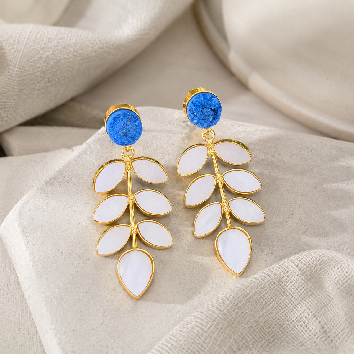 Angle view of gold-plated drop earrings with blue druzy and leaf-shaped mother of pearl details.