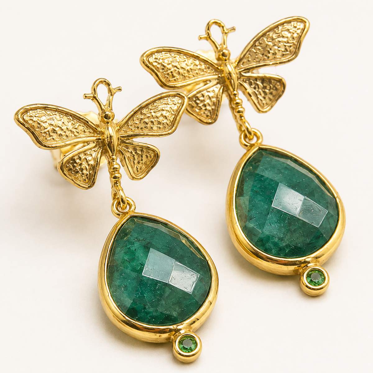 Close view of butterfly earrings with textured gold detail and green teardrop drops.