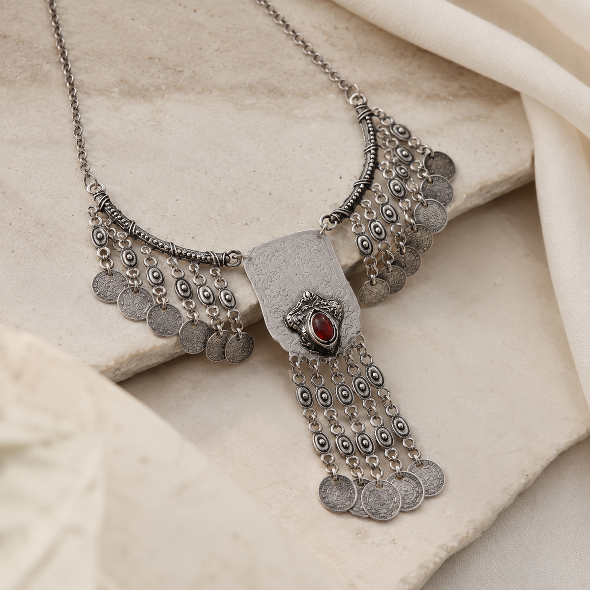 Angled view of a silver-tone coin fringe necklace with a red stone centerpiece.