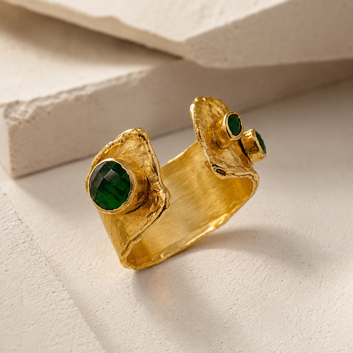 Angle view of a textured gold-plated open cuff ring with emerald green stone accents.