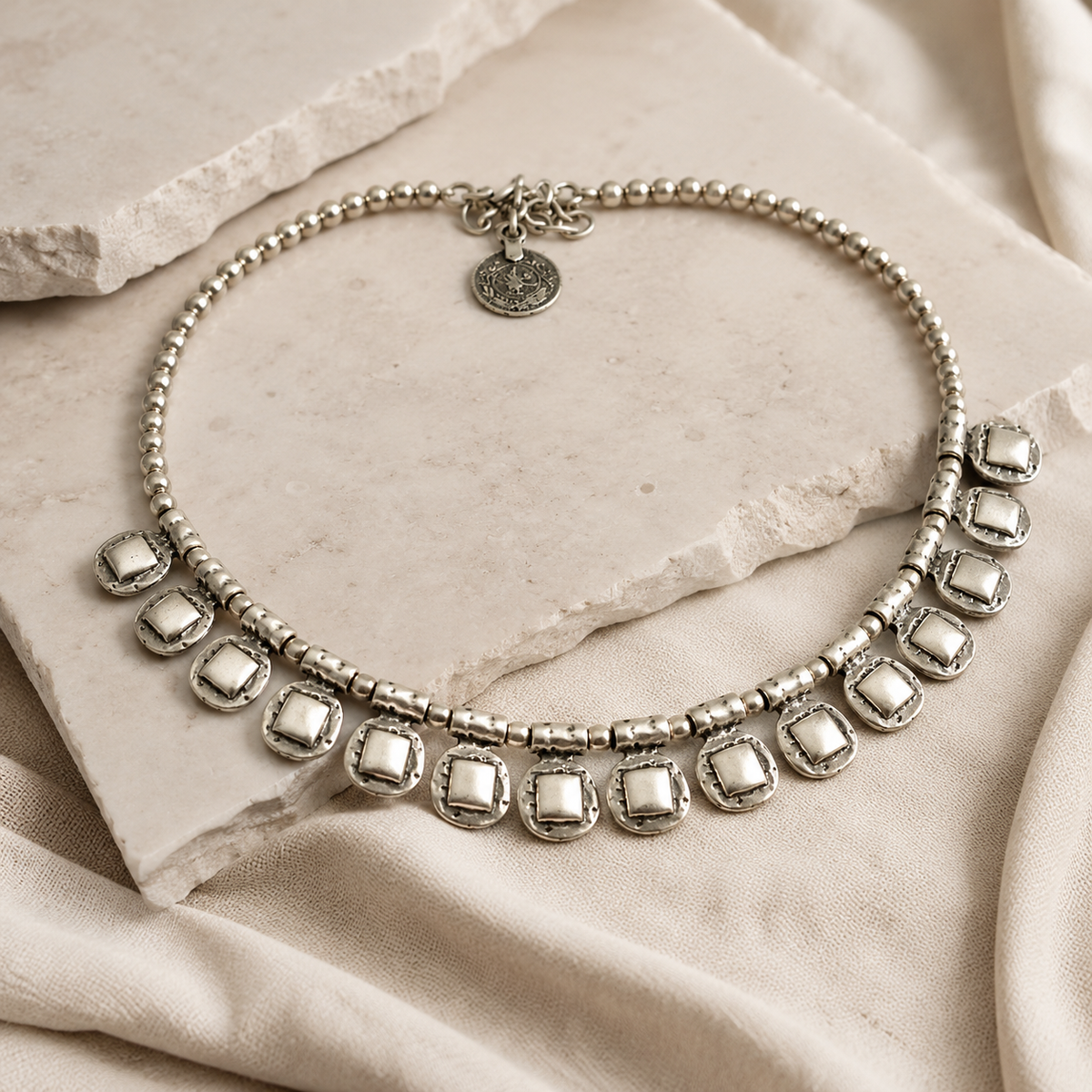 Angled view of a silver-tone beaded necklace with textured square drop accents.