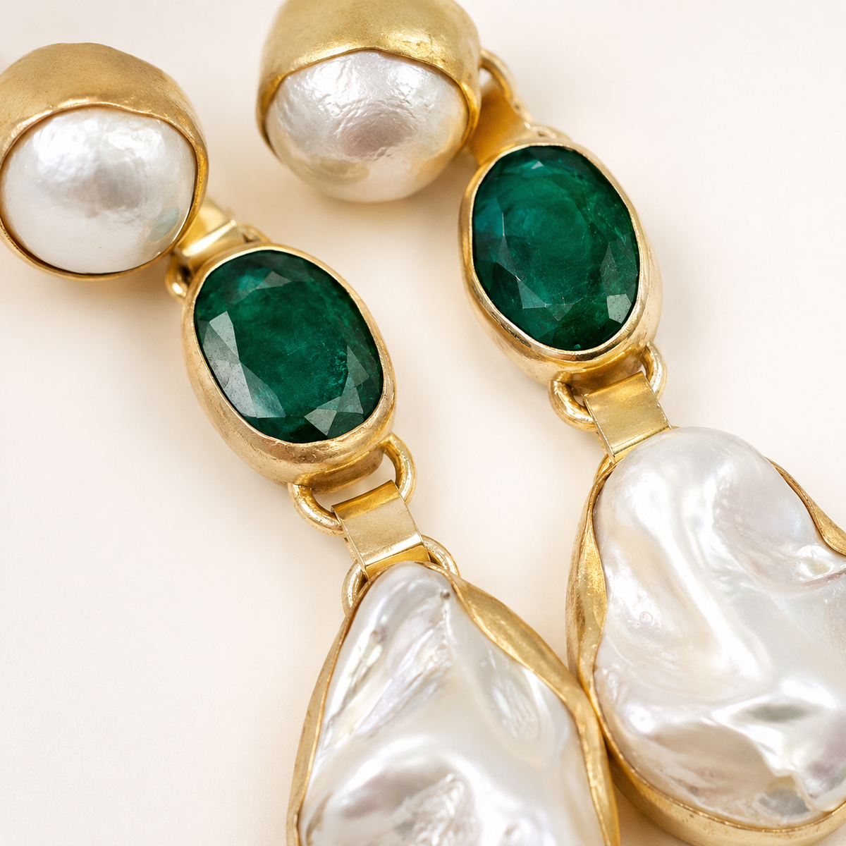 Close view of gold-plated earrings with emerald green oval stones and baroque pearl-style details.