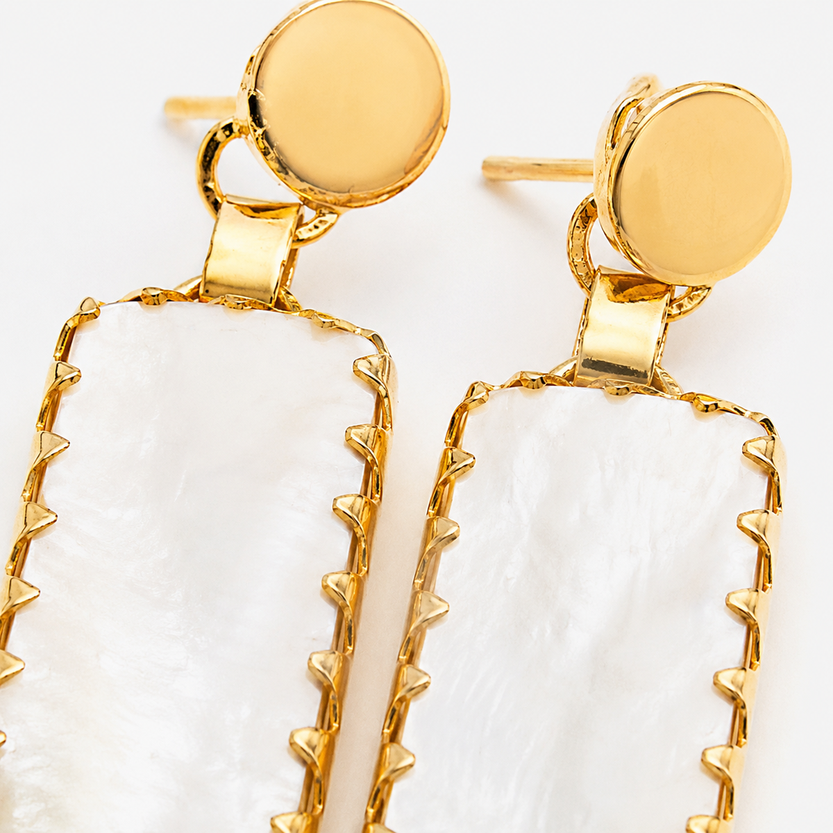 Close view of gold-plated earrings with textured edges and smooth pale green and white stone-like drops.