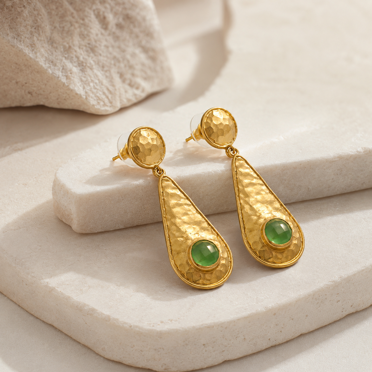 Angle view of hammered gold-plated drop earrings with green stone detail.