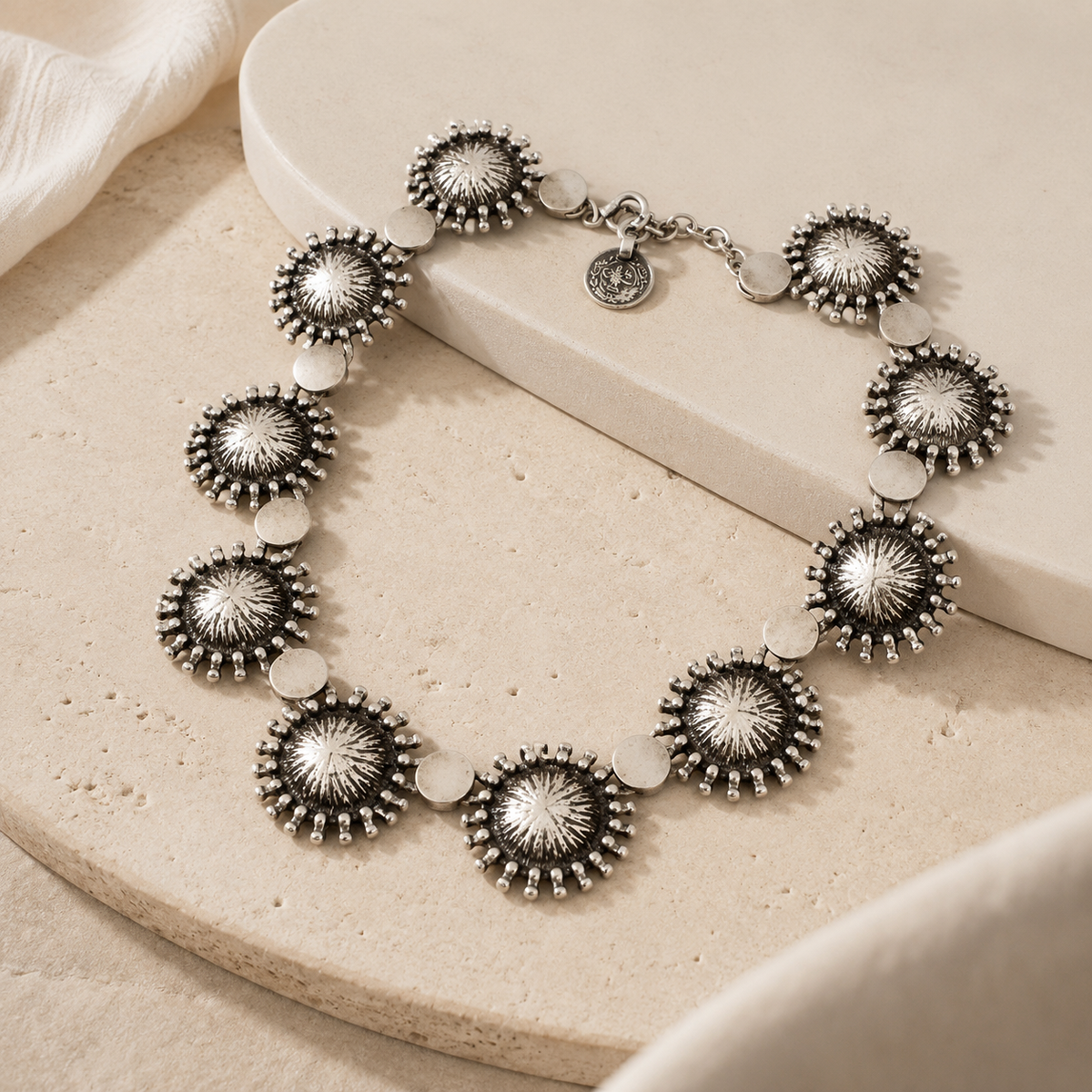 Angled view of a silver-tone sunburst disc collar necklace with textured details.