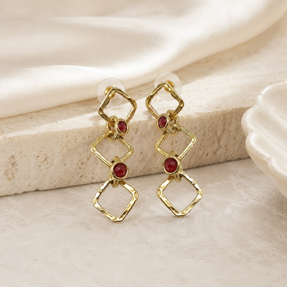Gold-tone drop earrings with a geometric chain design and red stone accents on a white background.