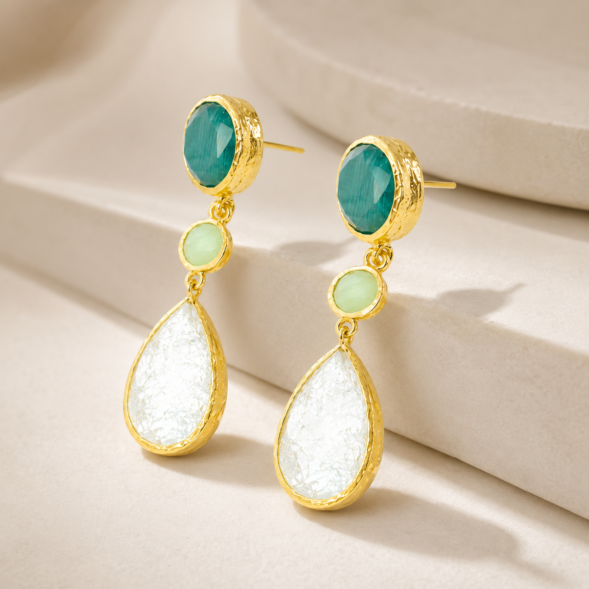 Angled view of gold-plated dangle earrings with faceted green and clear stones.