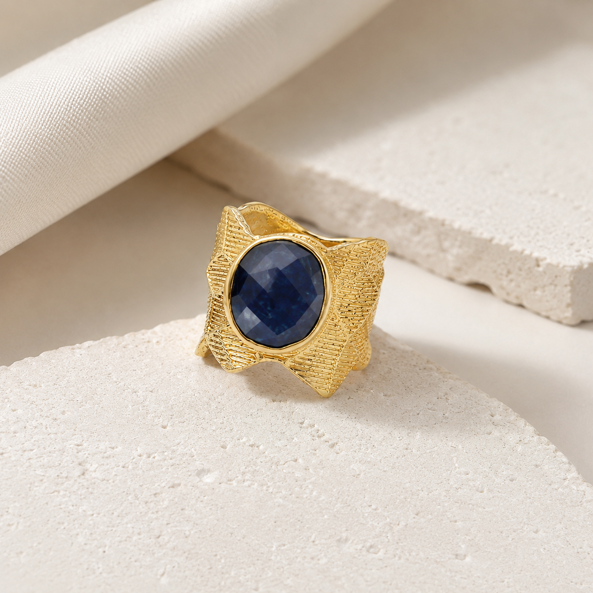 Angled view of a gold-plated ring with a textured geometric setting and navy blue oval stone.