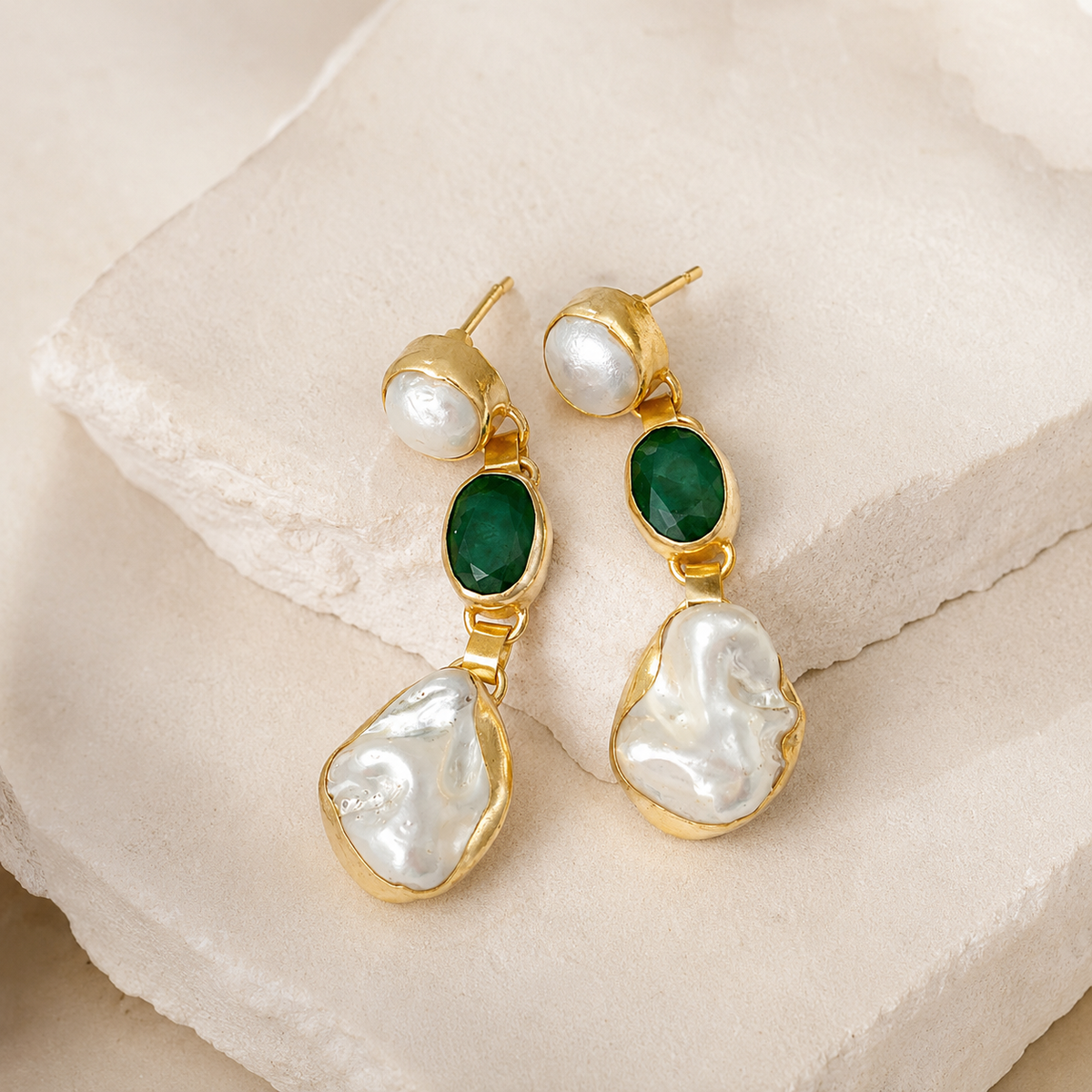 Angled view of gold-plated drop earrings with emerald green oval stones and baroque pearl-like drops.