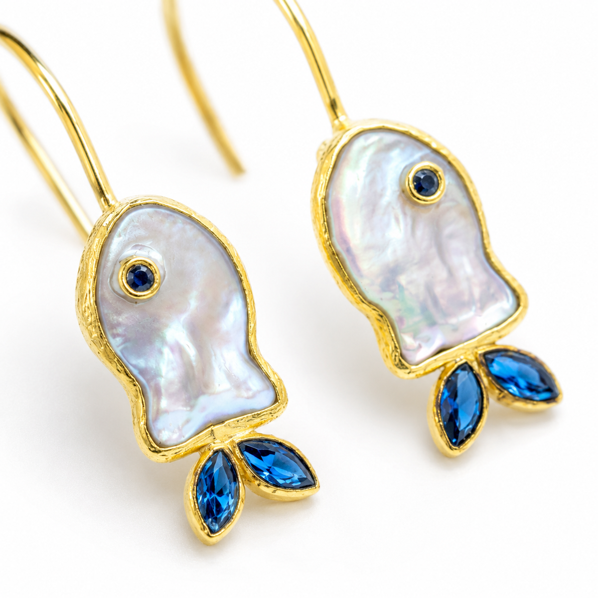 Close view of mother-of-pearl fish earrings with blue CZ details and gold-plated edges.