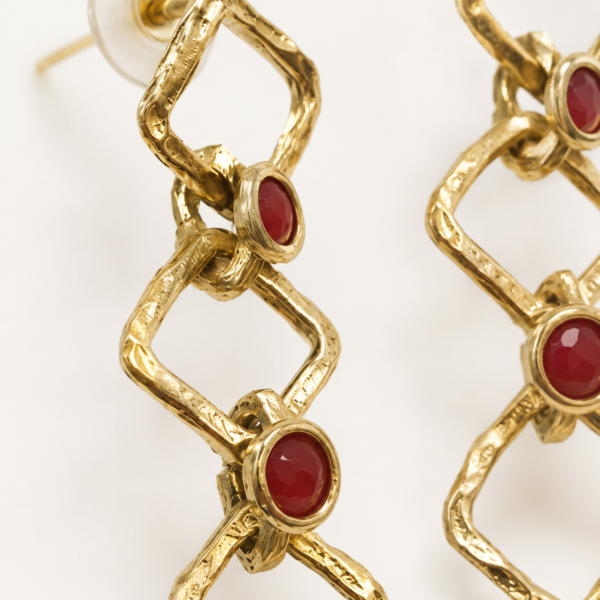 Close view of gold-tone linked earrings with textured edges and red stone details.