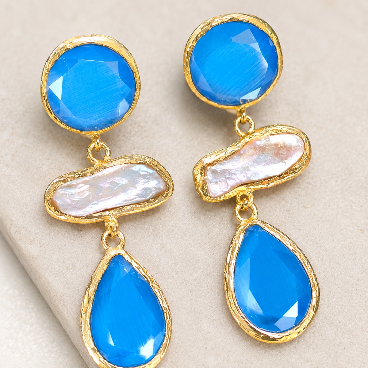 Close view of gold-plated earrings with blue faceted stones and a pearl-like center detail.