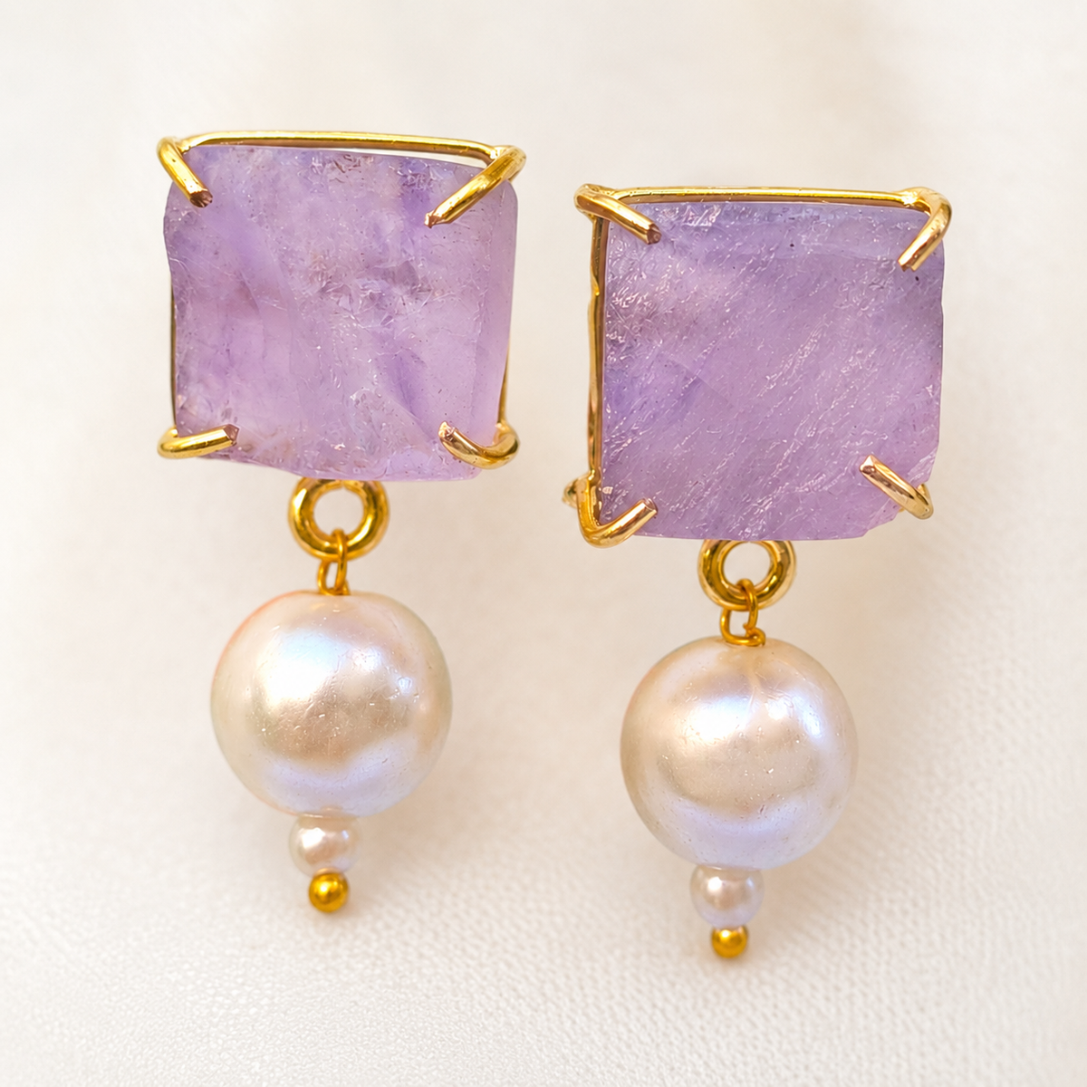 Close-up of lavender stone earrings with gold-plated prongs and pearl drops.