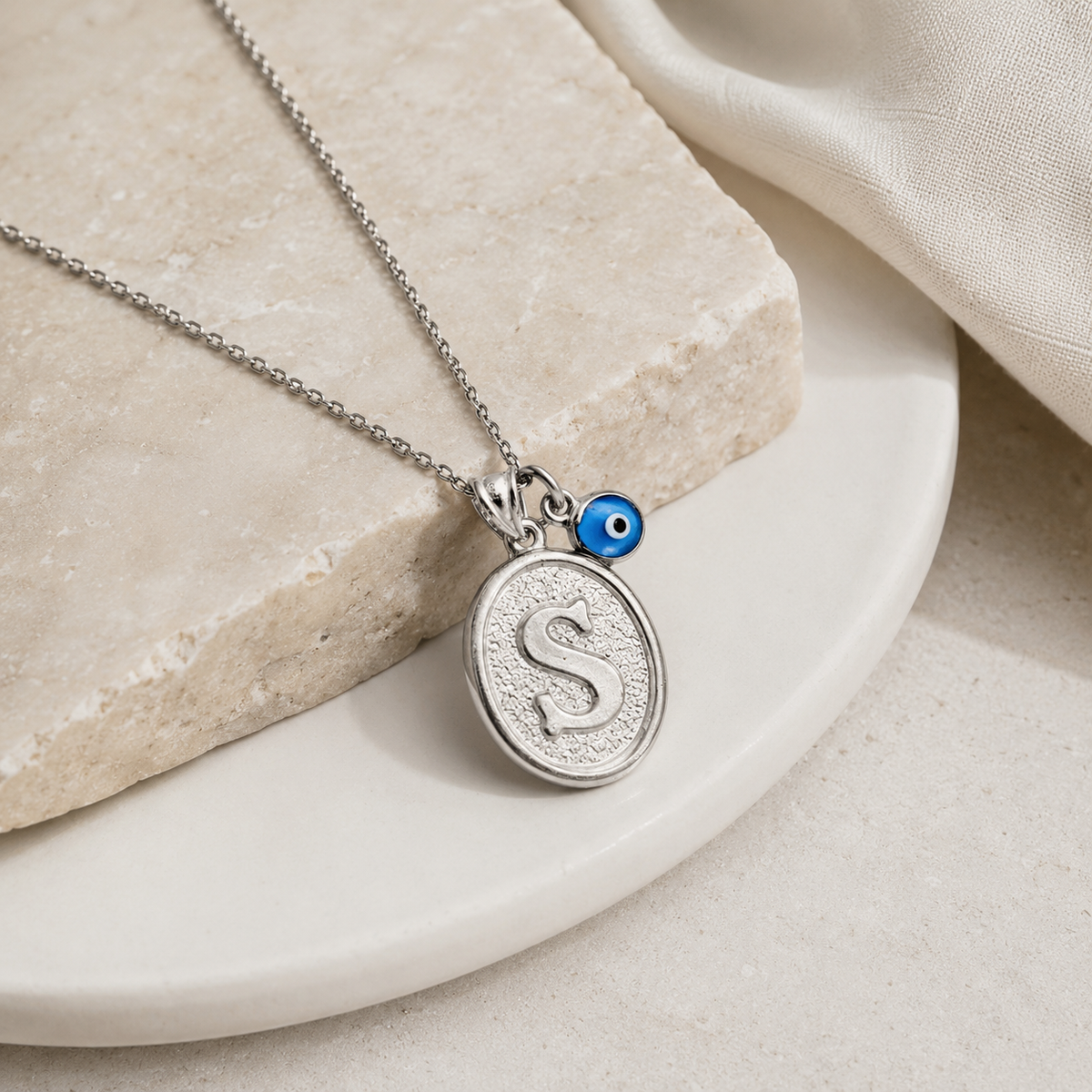 Silver-tone initial necklace with an oval pendant and blue eye charm on a neutral stone surface.