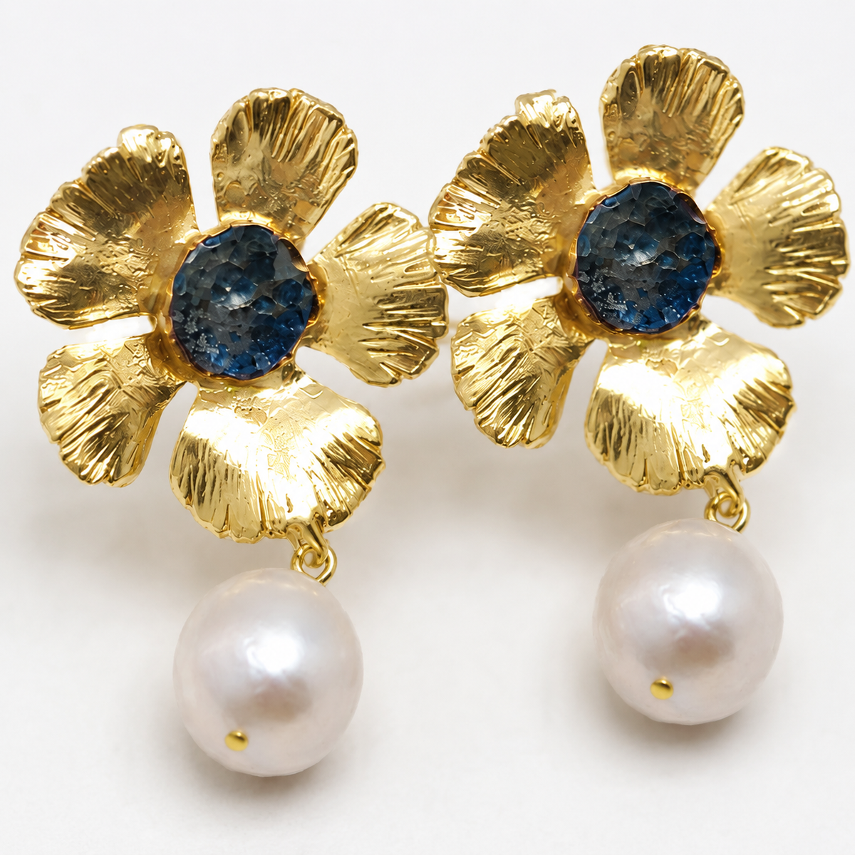 Close view of gold flower earrings with textured petals, a blue center, and pearl drop.