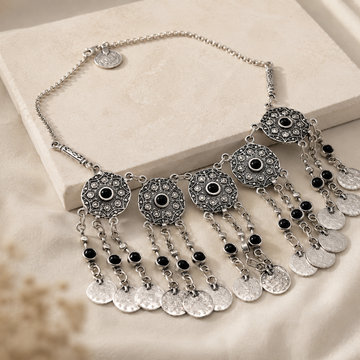 Angled view of a silver-tone statement necklace with coin fringe and black stone accents.